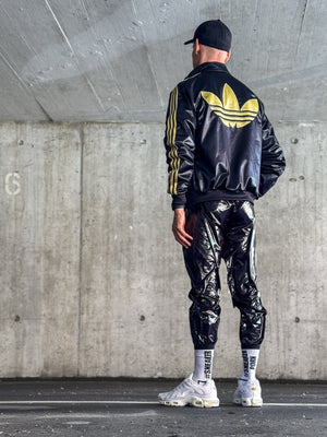 Eine Person steht unter einer Betonstruktur, den Blick abgewandt, mit einer schwarzen Kappe, einer glänzenden schwarzen Hose, einer schwarzen Jacke mit einem großen gelben Adidas-Logo, weißen Turnschuhen und Sk8erboy® #SNEAKERPORN Socken schwarz.