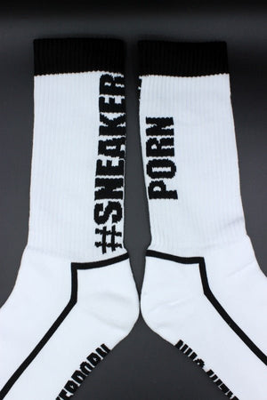 Ein Paar Sk8erboy® #SNEAKERPORN Socken schwarz mit schwarzen Zehen, Fersen und Bündchen zeigen die Worte "SNEAKER" und "PORN" in fetten schwarzen Buchstaben - echter Sneakerporn-Stil. Seite an Seite auf einer dunklen Oberfläche gelegt.