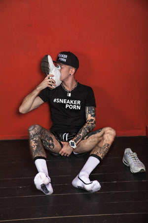 Eine tätowierte Person sitzt auf einem dunklen Holzboden an einer roten Wand, trägt ein schwarzes Sneakerporn-Shirt und eine Kappe und küsst einen weißen Sneaker, während sie Sk8erboy® #SNEAKERPORN Socken schwarz trägt, mit einem weiteren Sneaker daneben.