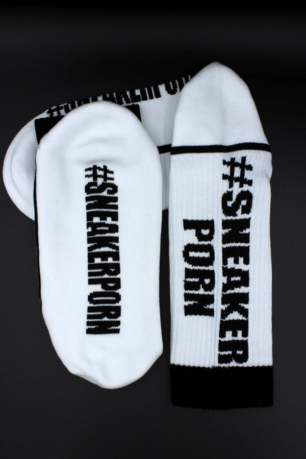 Ein Paar #SNEAKERPORN Socken schwarz von Sk8erboy® mit fettem schwarzem Text auf weißen Socken mit schwarzen Akzenten, perfekt für Sneakerporn-Fans und auf einem dunklen Hintergrund.