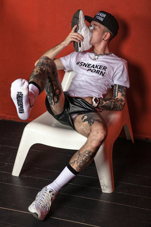 Eine tätowierte Person in einem weißen Sneakerporn-Shirt und einer Kappe sitzt auf einem weißen Stuhl vor einer roten Wand und schnüffelt an einem weißen Sneaker, während sie einen anderen mit schwarzen #SNEAKERPORN Socken von Sk8erboy® am Fuß trägt.